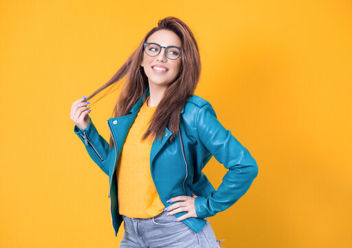 Happy young woman posing while playing with her hair, wearing blue leather jacket, isolated on yellow background. Playful cute girl