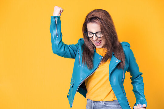 Excited beautiful young woman with closed eyes and clenched fists, wearing blue leather jacket, isolated on yellow background. Yes concept. Good news