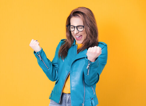 Excited young woman with clenched fists, wearing blue leather jacket, isolated on yellow background. Yes concept. Good news. Rejoicing girl celebrates success