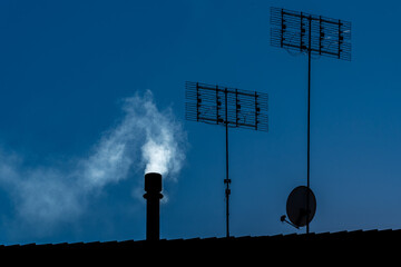 Chimney with white smoke, tv antennas and satellite dish seen in silhouette on rooftop, copy space
