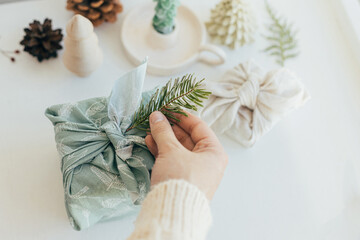 Zero waste Christmas concept. Hands decorating gift box wrapped in fabric with fir branch on white wooden table with eco friendly decor and candle. Furoshiki gift wrapping. Eco winter holiday