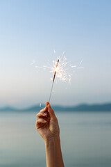A woman's hand holds burning sparklers against the backdrop of the sea and mountains close-up...