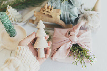 Hand holding wooden christmas tree toy on background of wrapped gifts in fabric, fir branch, candle on white table. Stylish eco friendly holiday decor and present. Merry Christmas and Happy Holidays