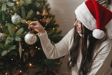 Happy woman in cozy sweater and santa hat decorating christmas tree with stylish baubles in...