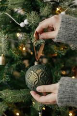 Hands decorating christmas tree with stylish bauble in atmospheric festive room. Merry Christmas! Winter holidays preparation. Woman in cozy sweater putting vintage toy on tree