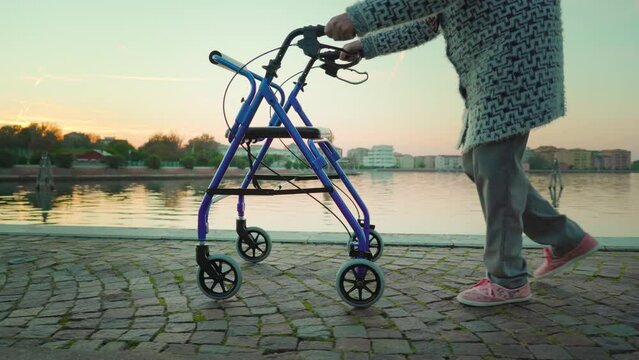 Old woman with walking frame wearing woolen cardigan and trousers strolls along road near Venice lagoon after sunset against Sottomarina