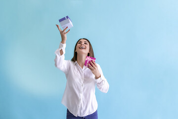 Portrait of happy beautiful brunette woman in white shirt catching falling pink and blue gift boxes against blue background. Selective focus. Holiday present theme.