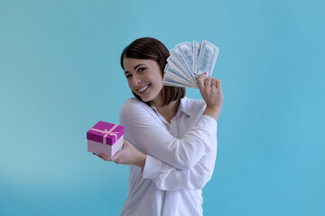 Portrait of beautiful smiling brunette woman holding pink gift box and stack of us dollars against blue background. Success theme.