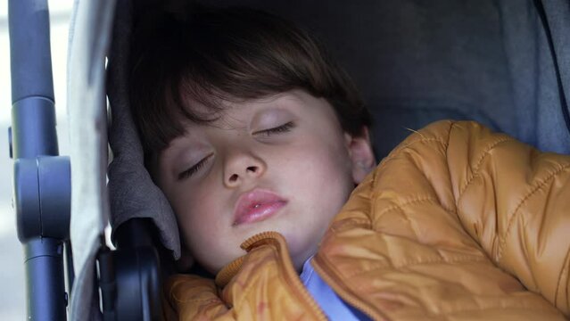 One little boy asleep sitting in stroller. Closeup child face sleeping wearing jacket. Kid napping in afternoon
