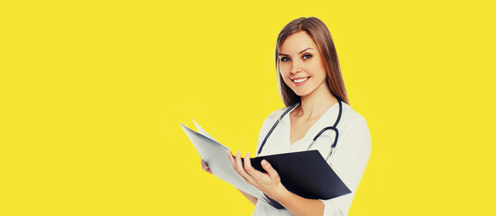 Portrait of friendly smiling young woman doctor with stethoscope and folder of documents on yellow background