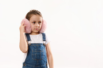 Beautiful Caucasian little child girl wearing blue denim overalls, cute pink plush headphones - winter earmuffs, ear warmers, isolated on white background. Protect your ears from cold. Copy space