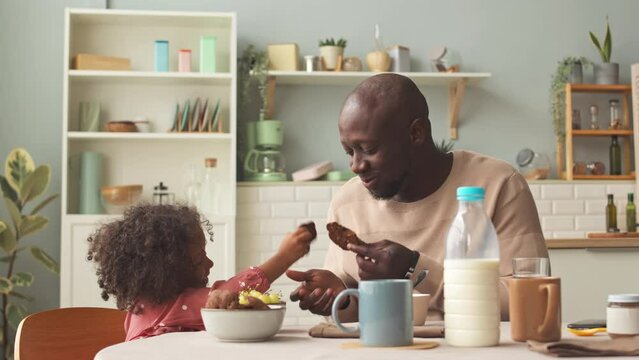 Cheerful Black man and his pretty little daughter sitting at kitchen table and having breakfast together