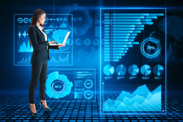 Attractive young european businesswoman using laptop with abstract glowing blue business charts and graphs hologram on dark background. Innovation, future and fintech concept.