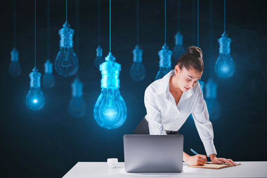 Attractive young european businesswoman leaning on desktop wigth laptop and writing with glowing digital lamp hologram on dark background. Idea, innovation and future concept.