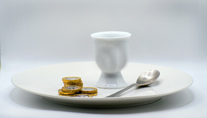 Dinner plate with British Currency pound coins and egg cup and spoon. Rising cost of food concept. Living with economising and budgeting for meals. Hunger.  Isolated white background with copy space. 