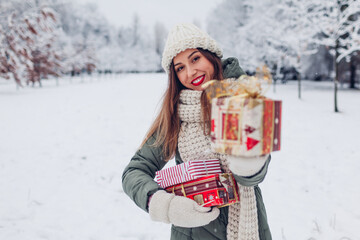Happy woman holding Christmas presents gift boxes in snowy winter park offering one. Festive holiday season. Space