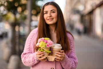 Fototapeta premium handsome woman gently holds flower arrangement in small pot and paper cup of coffee in her hands.