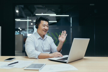 Working online meeting. Young handsome male Asian businessman conducts remote conference with partners. He sits in the office at the table with a laptop and headphones, greets the camera.