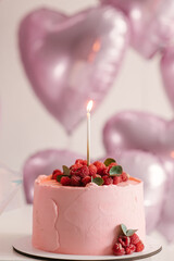 Festive pink cake with raspberries and a candle on the background of helium balloons of pink hearts