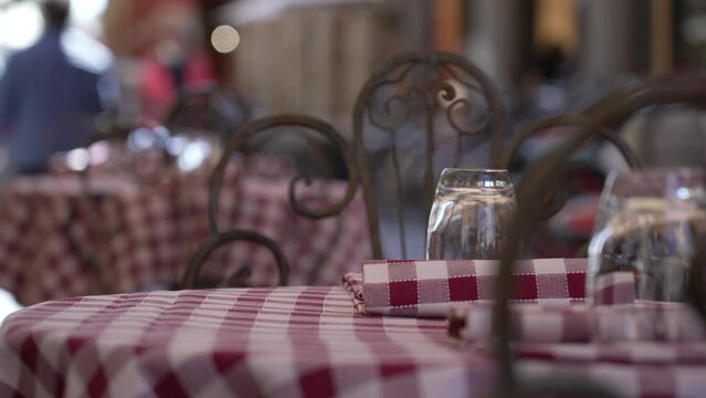 Empty Italian restaurant table in European street with napkin and glasses. tourists walking in background. Table Linens Checkpoint. A classic gingham print