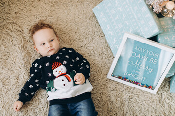 Naklejka premium A little curly boy in a traditional Christmas sweater spends time at home near the Christmas tree