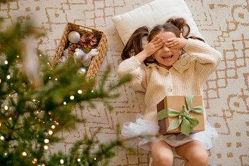 Fototapeta na wymiar A funny girl is lying under a Christmas tree with a gift box on the carpet, covering her eyes with her hands. The child rejoices at the advent of xmas