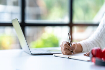 Cropped shot of Asian woman hand using laptop and writing making list taking notes in notepad working or learning on laptop  educational course or training, education online concept