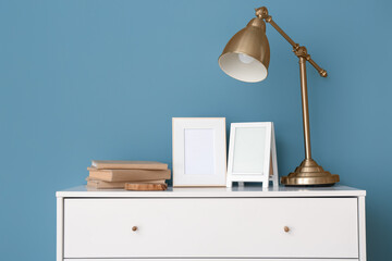 Blank frames, lamp and books on chest of drawers near blue wall