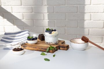 Yogurt in a glass jar with granola, chocolate, blueberries and honey on a wooden board on a white brick wall background with mint leaves. Front view