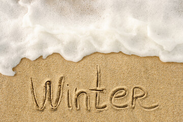 Fototapeta premium Winter word handwritten lettering on the sandy beach with white ocean wave background, seasonal concept photo