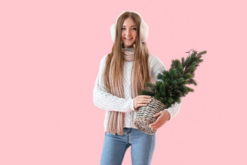 Young woman in warm ear muffs with small Christmas tree on pink background