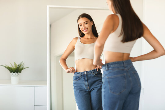 Cheerful young woman wearing jeans and smiling to her reflection in mirror after successful weight loss, copy space