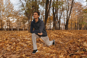 Fit male runner in sportswear doing lunge exercise and stretching legs while training in nature in autumn  - obrazy, fototapety, plakaty