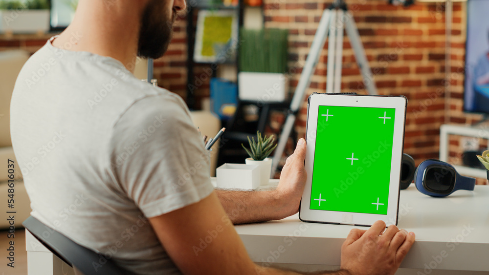 Poster young man vertically holding digital tablet with greenscreen template in living room, sitting at des
