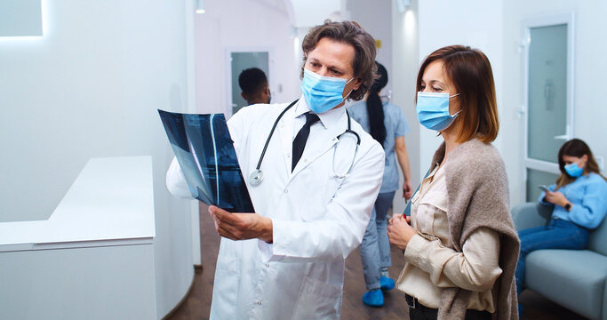 Portrait Of Caucasian Male Physician In Medical Mask Standing In Clinic And Talking To Female Patient Discussing X-ray Scan. Multi-ethnic Healthcare Workers Speak On Background. Covid-19 Concept