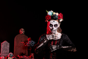 Fototapeta premium Horror lady goddess of death costume with body art, wearing flowers crown to celebrate dios de los muertos tranditional mexican holiday. Woman with santa muerte skull make up in studio.