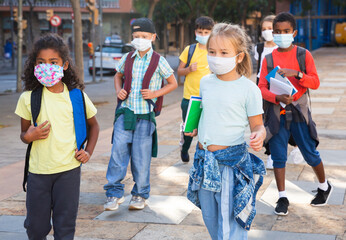 Multiethnic group of tweenagers in medical masks walking outside school building on autumn day, going to lessons. Concept of necessary .precautions in COVID pandemic..