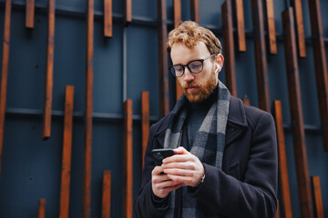 A young red-haired guy, a businessman in glasses, against the backdrop of urban style, checks mail on a smartphone, communicates in a messenger and drinks coffee. Online communication.
