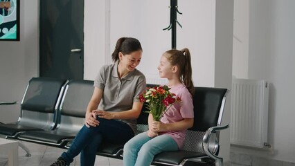 Mother and little child sitting in waiting room before medical appointment, girl holding bouquet of flowers. Young kid and woman waiting to attend examination with physician in lobby.