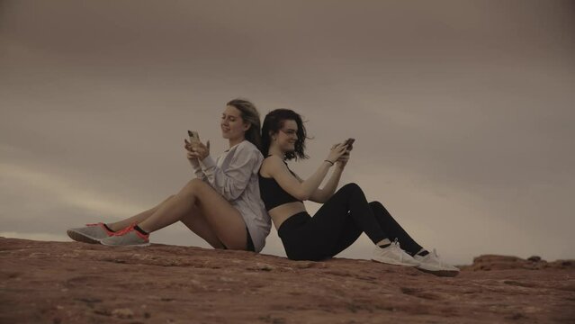 Slow motion of girls sitting back to back in desert posing for cell phone selfies / St. George, Utah, United States