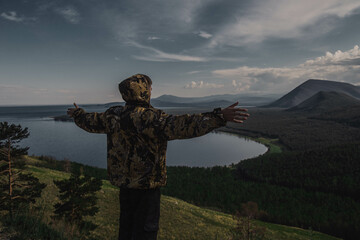 a young man in a khaki jacket stands with his back, arms outstretched, mountains