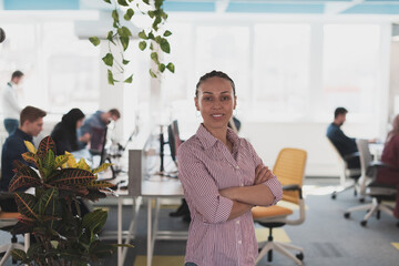 Portrait of young smiling business woman in creative open space coworking startup office. Successful businesswoman standing in office with copyspace. Coworkers working in background.