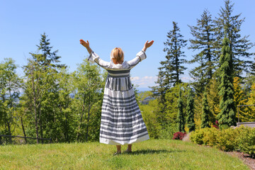 A woman in a white dress with white hair stands facing the forest with her hands up, praise and delight