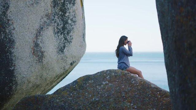 Photographer, beach and peace with a woman in nature, sitting on a rock boulder to enjoy the ocean view of the horizon. Water, travel and freedom with a female by the sea to relax on summer vacation