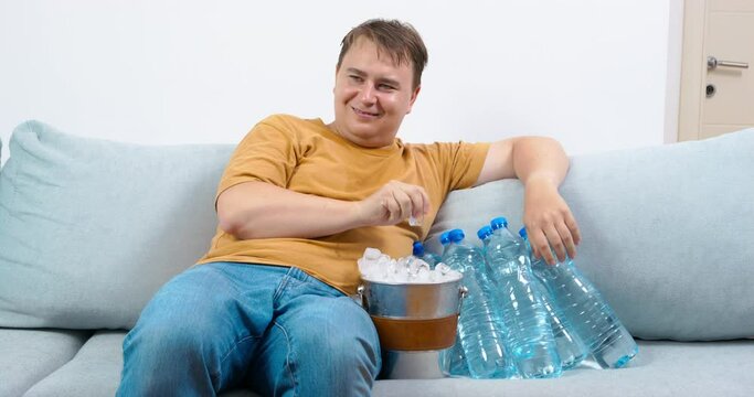 Mature plump man sits on comfortable sofa near bottles of water and bucket filled with ice. Man picks out ice cubes on hot summer day looking satisfied