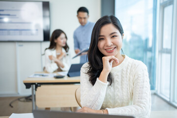 Obraz premium Happy beautiful Asian businesswoman sitting smiling and enjoying working on laptop in office and looking at camera.