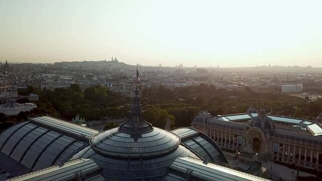 Drone tour of Paris city from above, with French flag waving on top of Grand Palais and Petit Palais at the dawn, highlighting silhouette view of Sacre Coeur at distance.