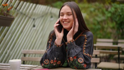Charming young woman with toothy smile talking on phone sitting outdoors. Portrait of happy beautiful lady in national Ukrainian embroidered shirt chatting on smartphone in slow motion smiling
