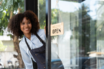 Small business african female owner smiling while turning sign for opening of cafe. Happy afro-american waitress entrepreneur in apron present sign on door