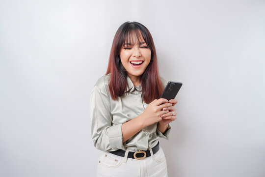 A portrait of a happy Asian woman wearing a sage green shirt and holding her phone, isolated by white background